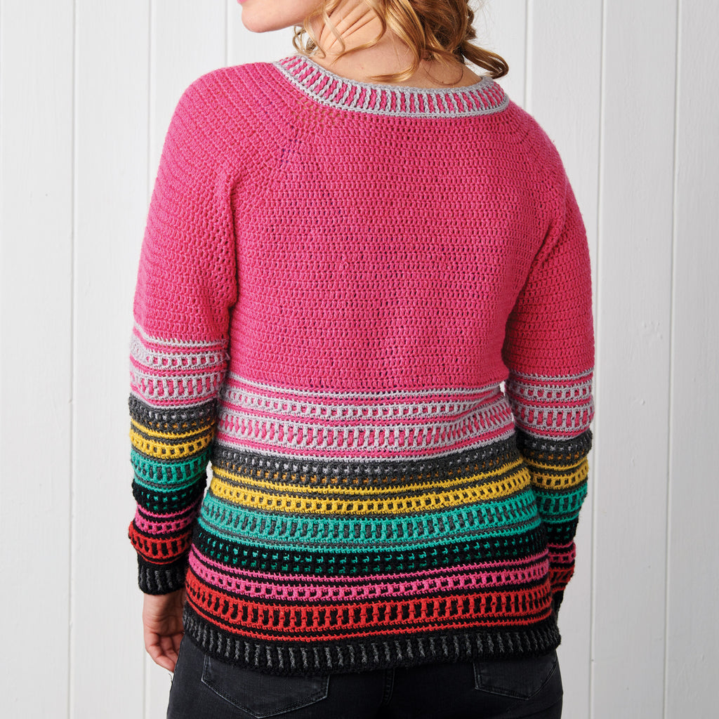Woman wearing a colorful crochet sweater. The top is pink, with multicolored horizontal stripes below in gray, yellow, teal, and red hues. White wooden background.
