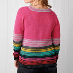 Woman wearing a colorful crochet sweater. The top is pink, with multicolored horizontal stripes below in gray, yellow, teal, and red hues. White wooden background.