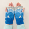 A pair of knitted wristwarmers with a mountain and patchwork pattern in blue and white colors, being worn by a person.