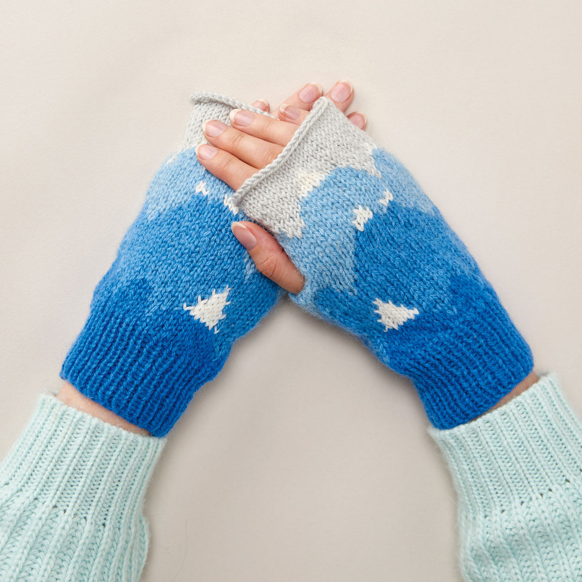 A pair of knitted wristwarmers with a mountain and patchwork pattern in blue and white colors, being worn by a person.