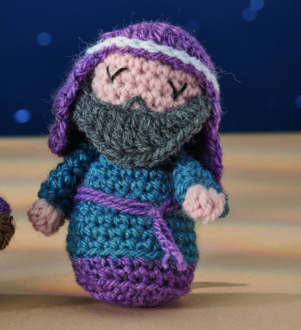 Crocheted doll with a gray beard, wearing a blue and purple robe and headdress, set against a deep blue background with a serene expression.