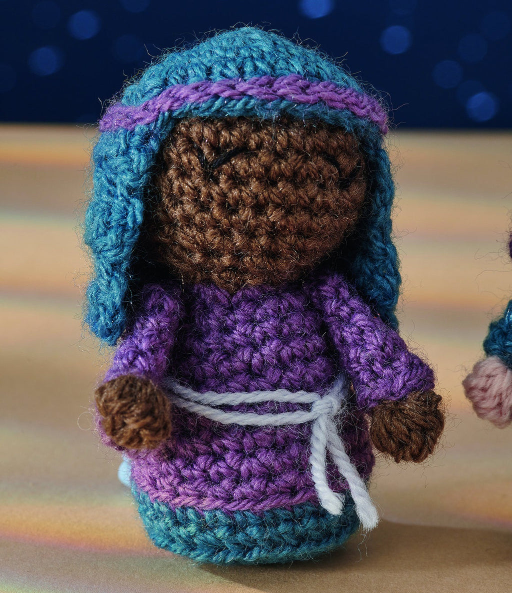 Crocheted doll with a brown face, smiling eyes, wearing a blue headdress, purple robe, and white belt. Soft focus background with blue lights.