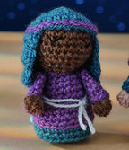 Crocheted doll with a brown face, smiling eyes, wearing a blue headdress, purple robe, and white belt. Soft focus background with blue lights.