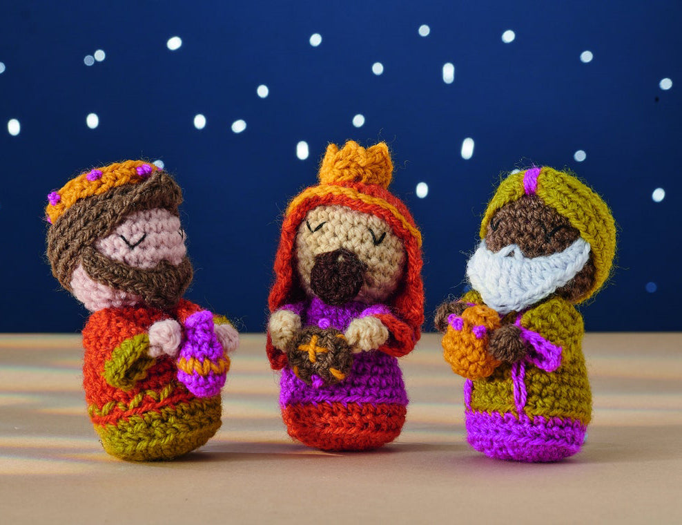 Three colorful crochet figures of the Three Wise Men, each holding gifts, against a dark blue, starry backdrop, conveying a whimsical and festive tone.