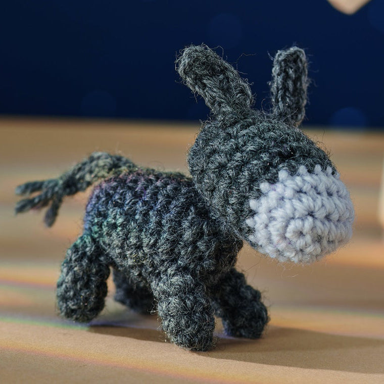 A crocheted donkey toy in dark, textured yarn stands on a smooth, brown surface. Its face is light gray, set against a blurred blue background.