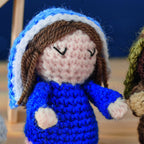 Crocheted doll in blue and white clothing, featuring brown yarn hair and closed eyes. The background is softly blurred, creating a cozy, handmade feel.