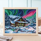 Framed cross-stitch art of a snowy cabin with Northern Lights