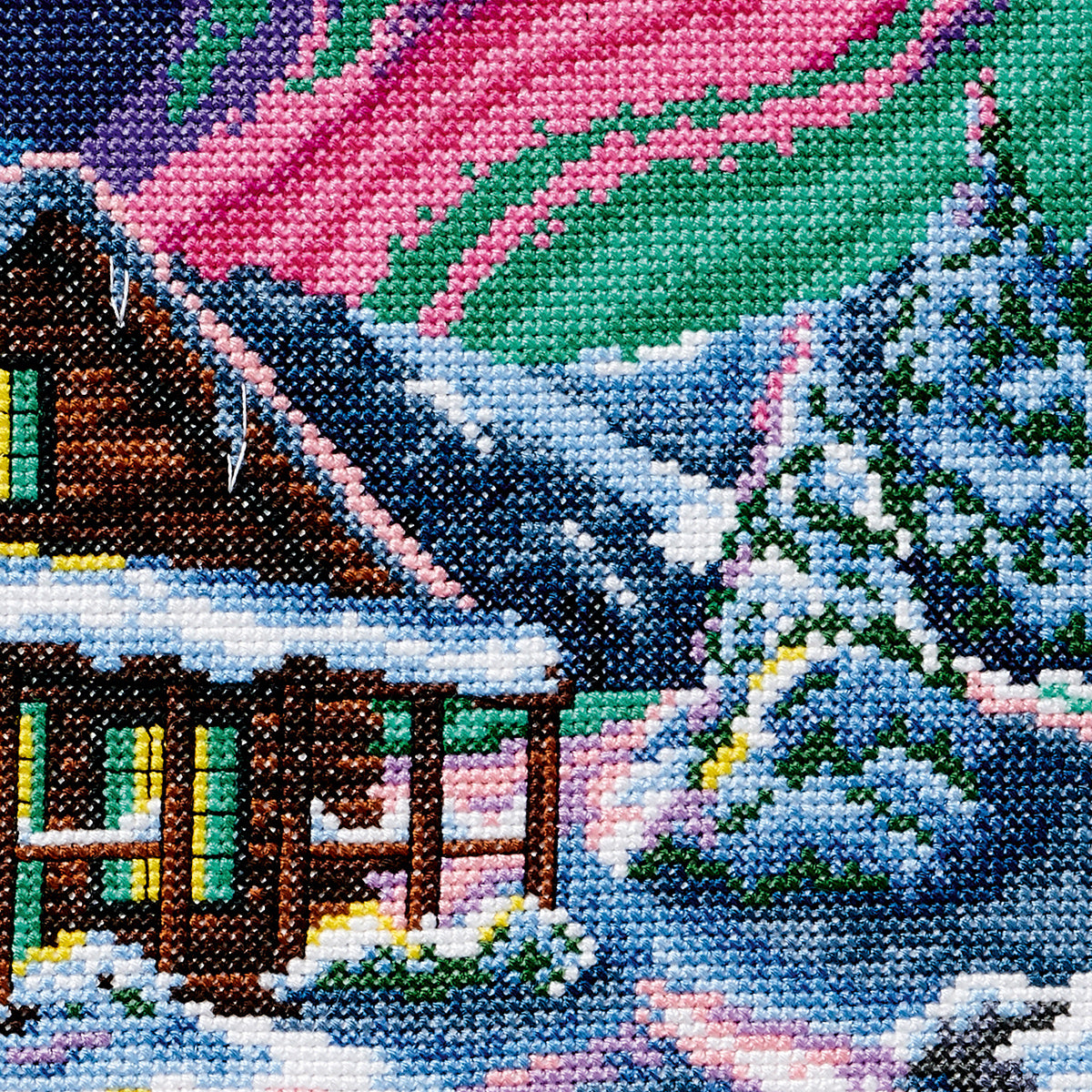 Cross-stitched scene of a snowy cabin with trees and a colorful sky.