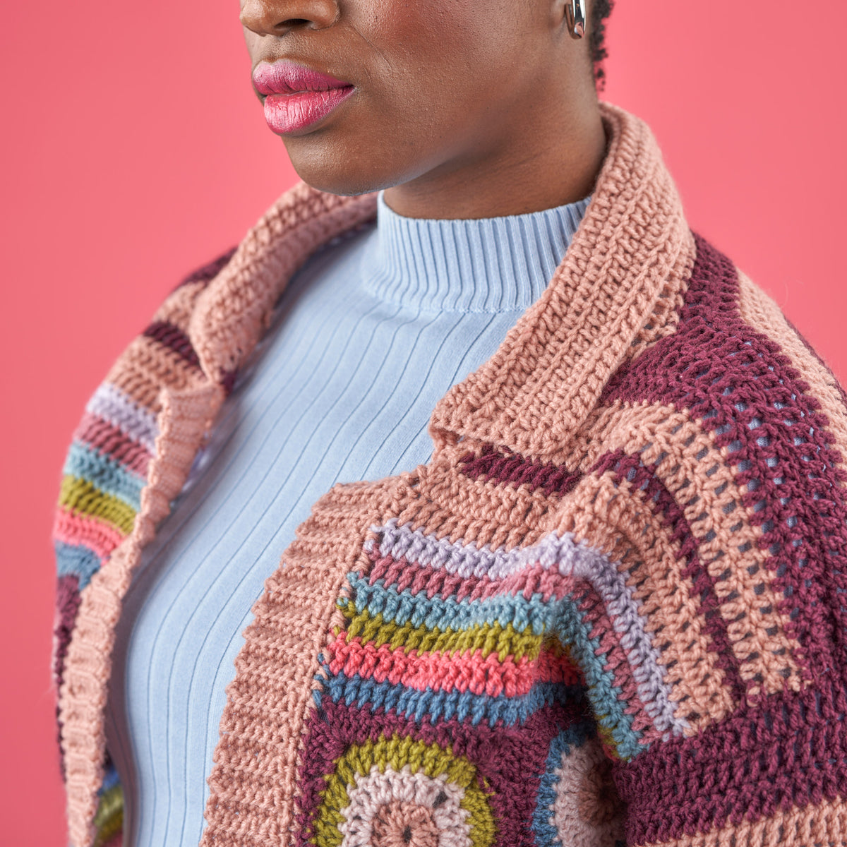Person wearing a colorful crocheted cardigan against a pink background