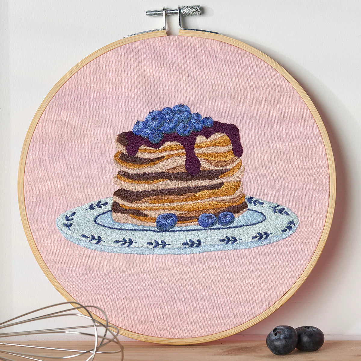 Embroidery hoop with a stack of pancakes and blueberries on a pink background