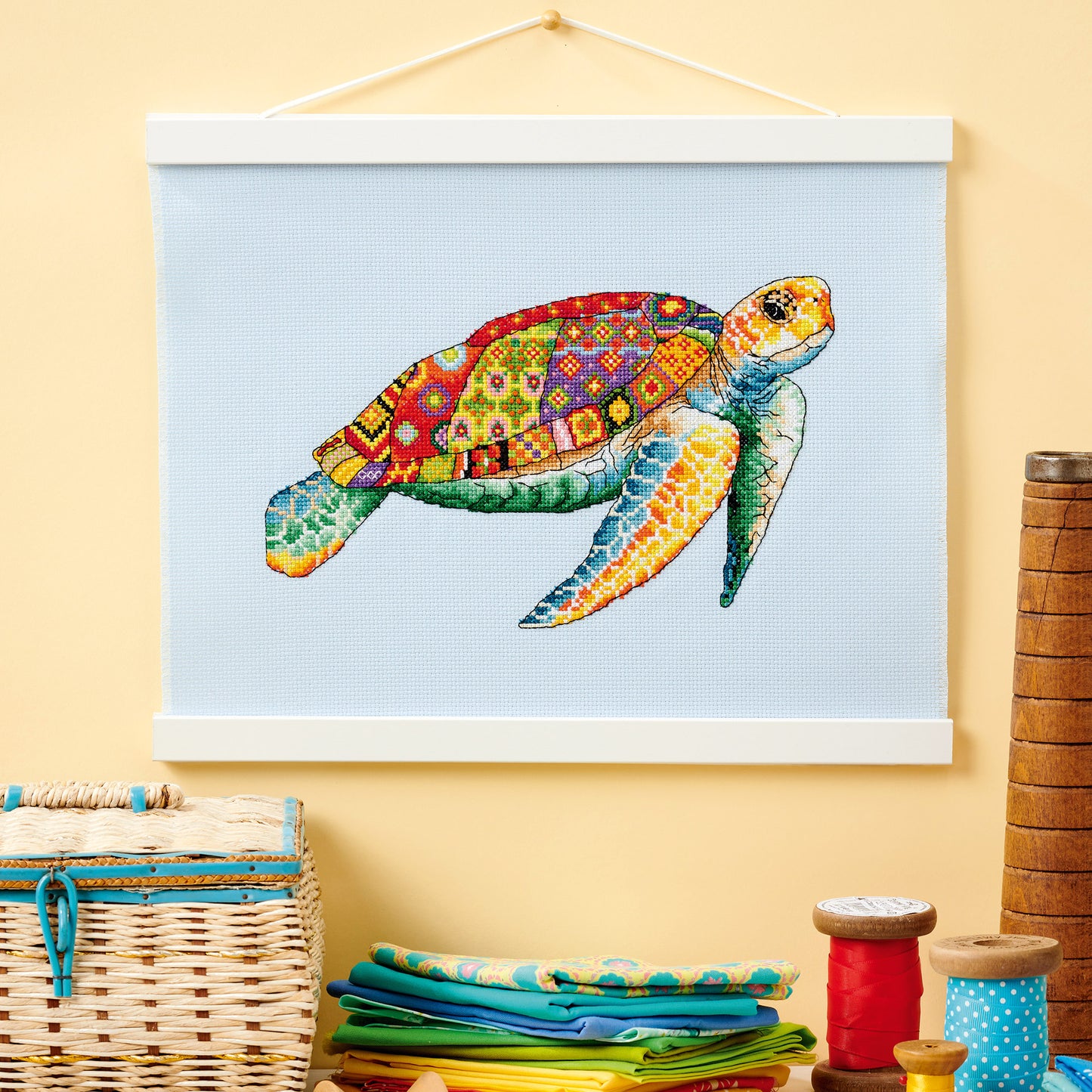 Colorful turtle cross-stitch pattern on a light blue background, hanging on a yellow wall with sewing supplies below.