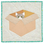 Quilt block with a cat peeking out from a cardboard box on a woven background