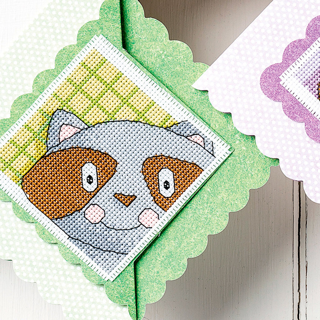 Cross-stitched design of a raccoon face on green fabric with a scalloped edge.