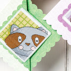 Cross-stitched design of a raccoon face on green fabric with a scalloped edge.