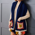 Person wearing a blue crochet vest with colorful square patterns and pom-poms against a gray background