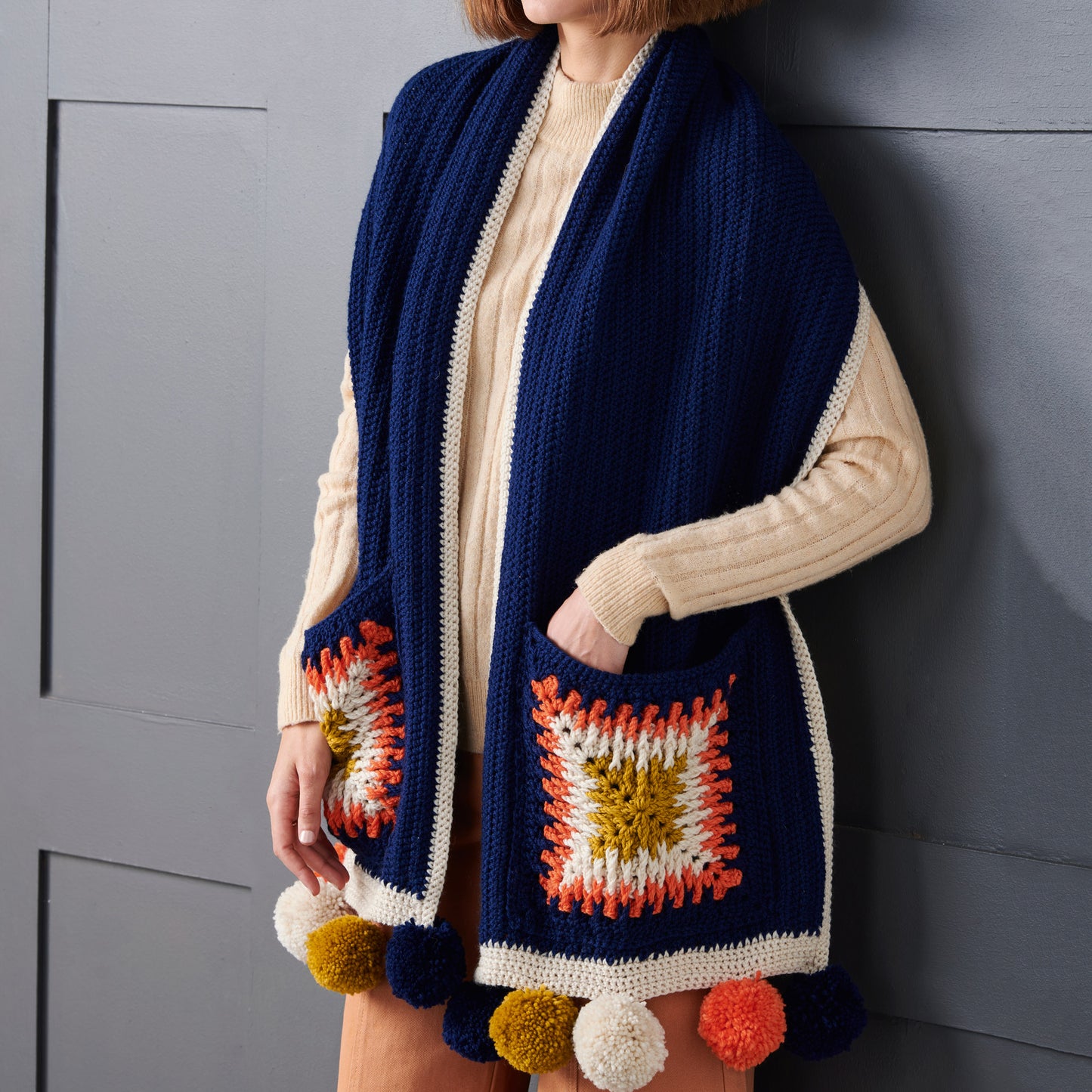 Person wearing a blue crochet vest with colorful square patterns and pom-poms against a gray background