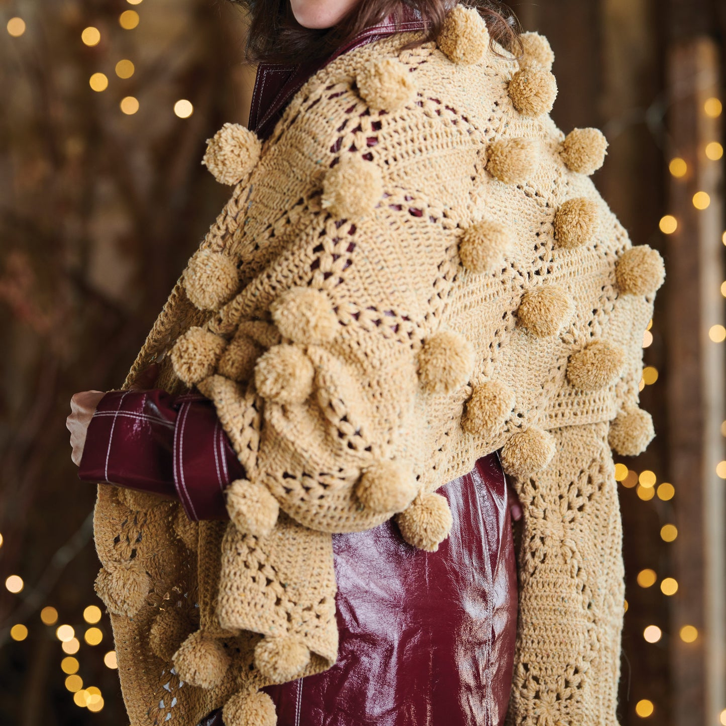 Person wearing a beige crochet shawl with pom-poms against a blurred background with lights.