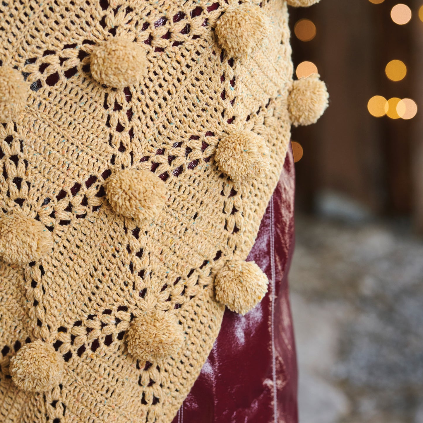 Close-up of a beige crochet shawl with pom-poms against a blurred background
