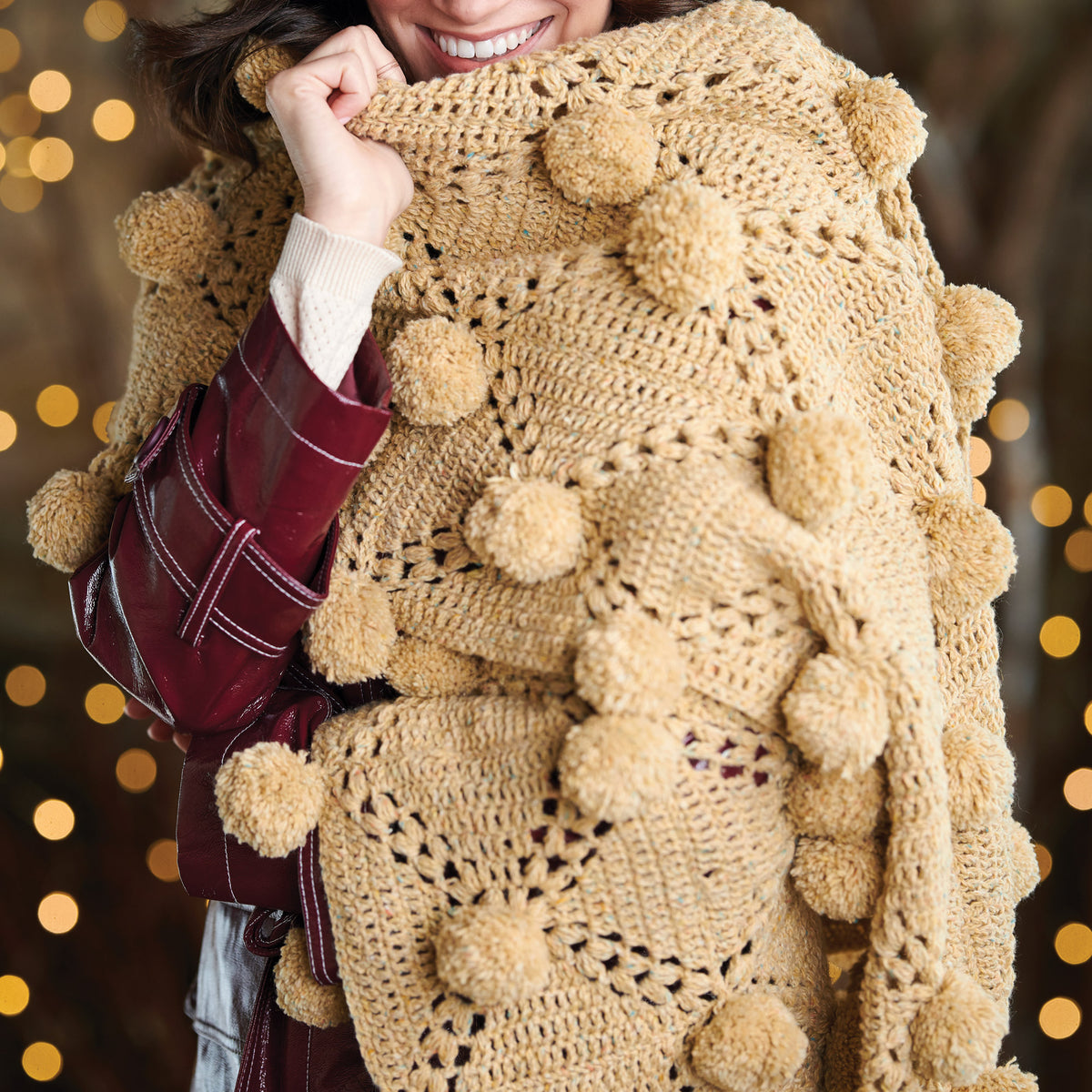 Person wearing a beige crochet sweater with pom-poms against a blurred background with lights.