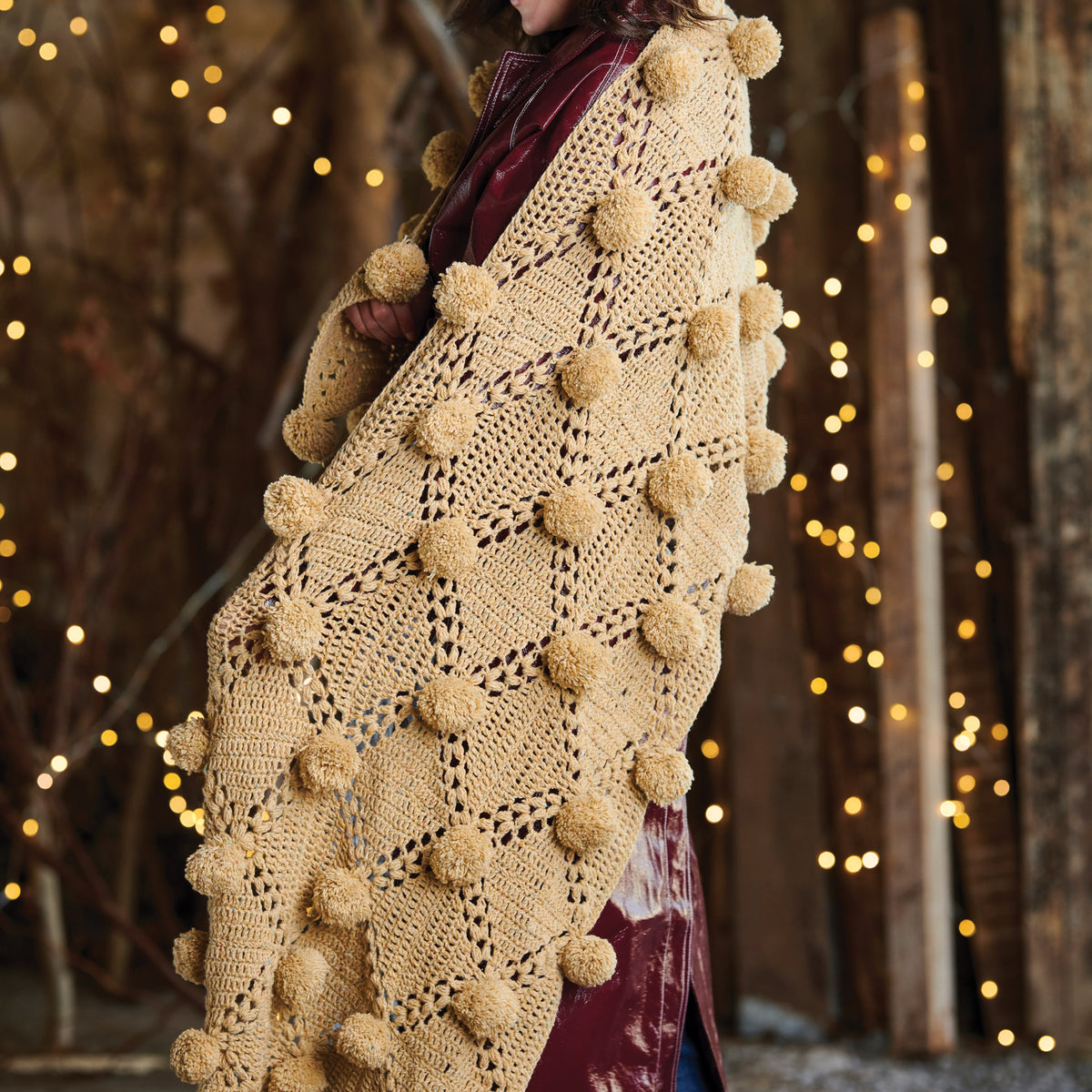 Beige crochet shawl with pom-poms worn by a person against a rustic background with lights.