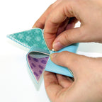 Hands folding a blue and green paper triangle with a purple pattern on a white background