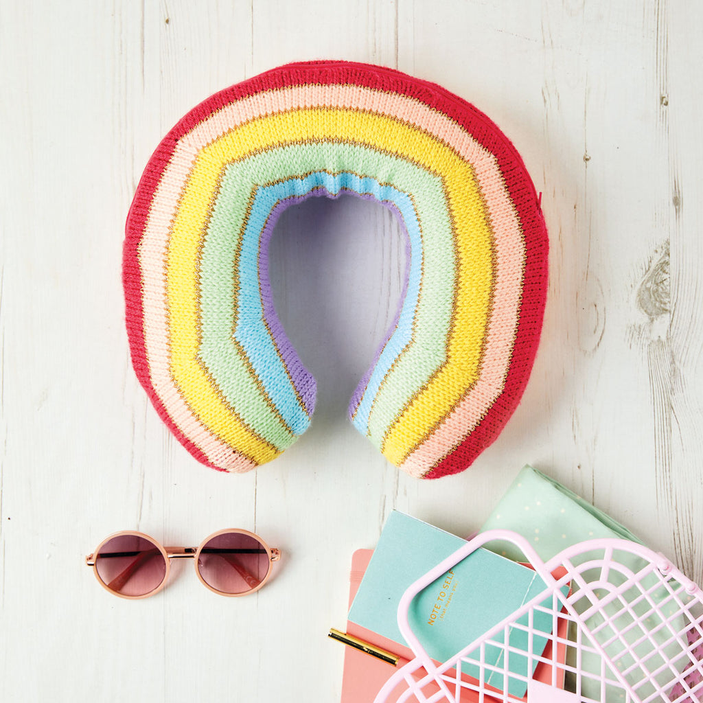 A knitted cushion in a rainbow stripe pattern, displayed on a wooden surface accompanied by sunglasses and a notebook.
