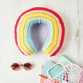 A knitted cushion in a rainbow stripe pattern, displayed on a wooden surface accompanied by sunglasses and a notebook.