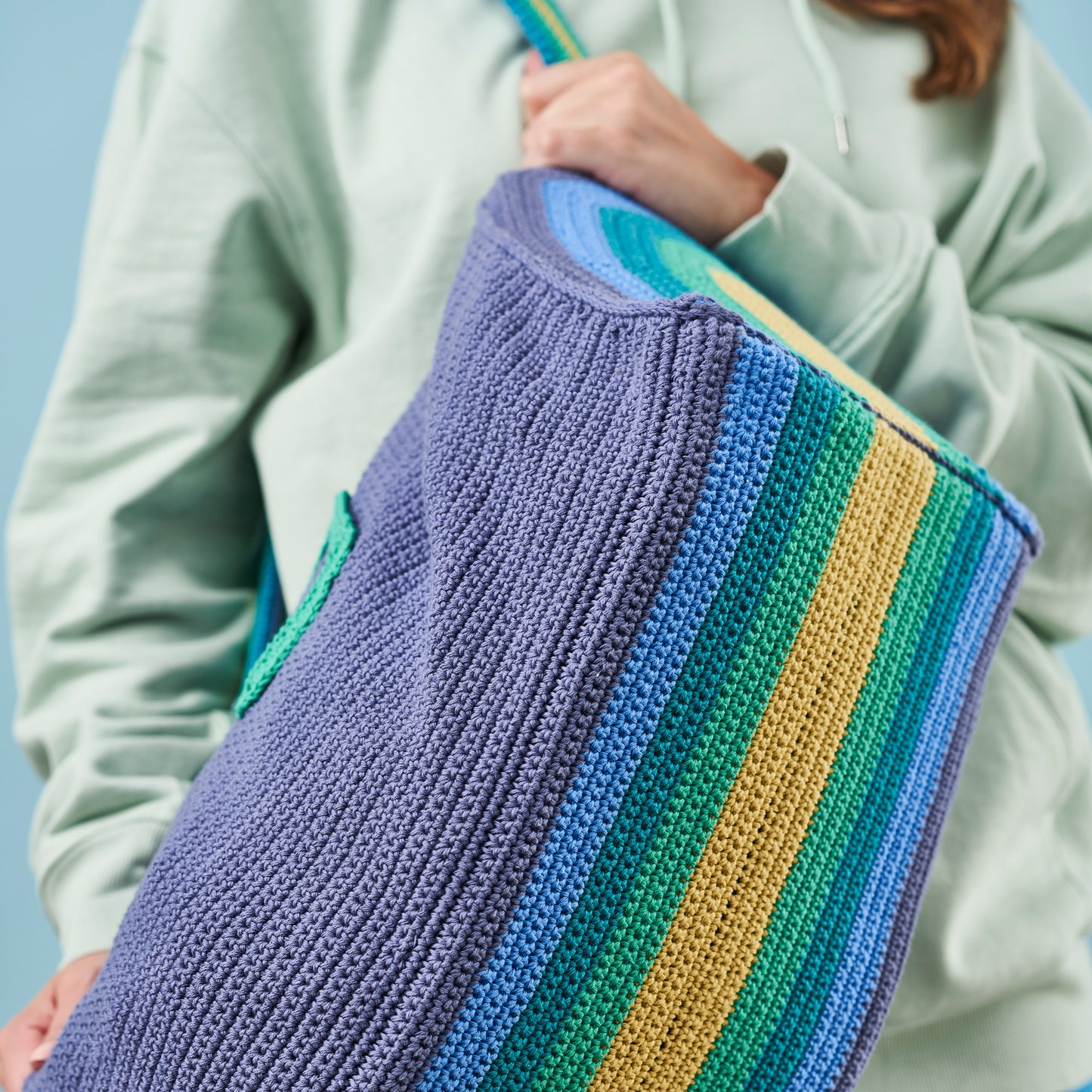 Person holding a colorful striped crochet blanket against a light blue background