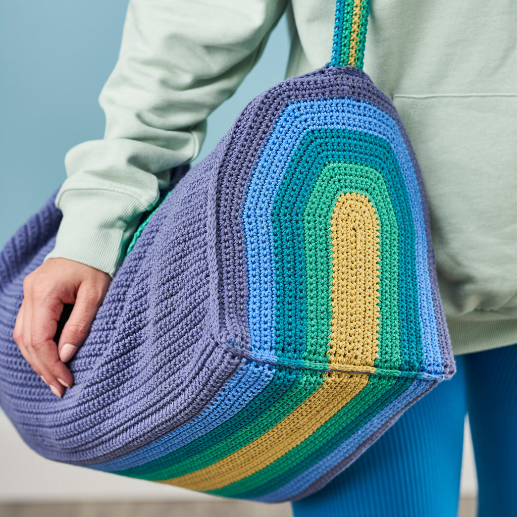 Person holding a colorful crocheted bag with a gradient of blue, green, and yellow.