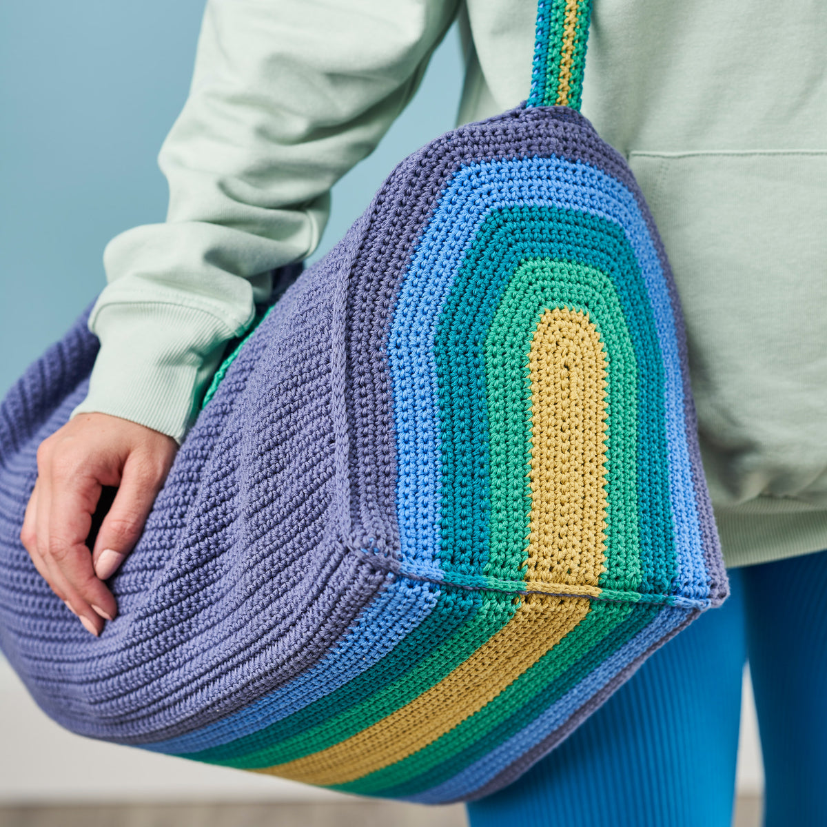 Person holding a colorful crocheted bag with a gradient of blue, green, and yellow.