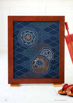Framed textile art with geometric patterns and circular motifs in white on a dark blue background. A red umbrella and traditional sandals are nearby.