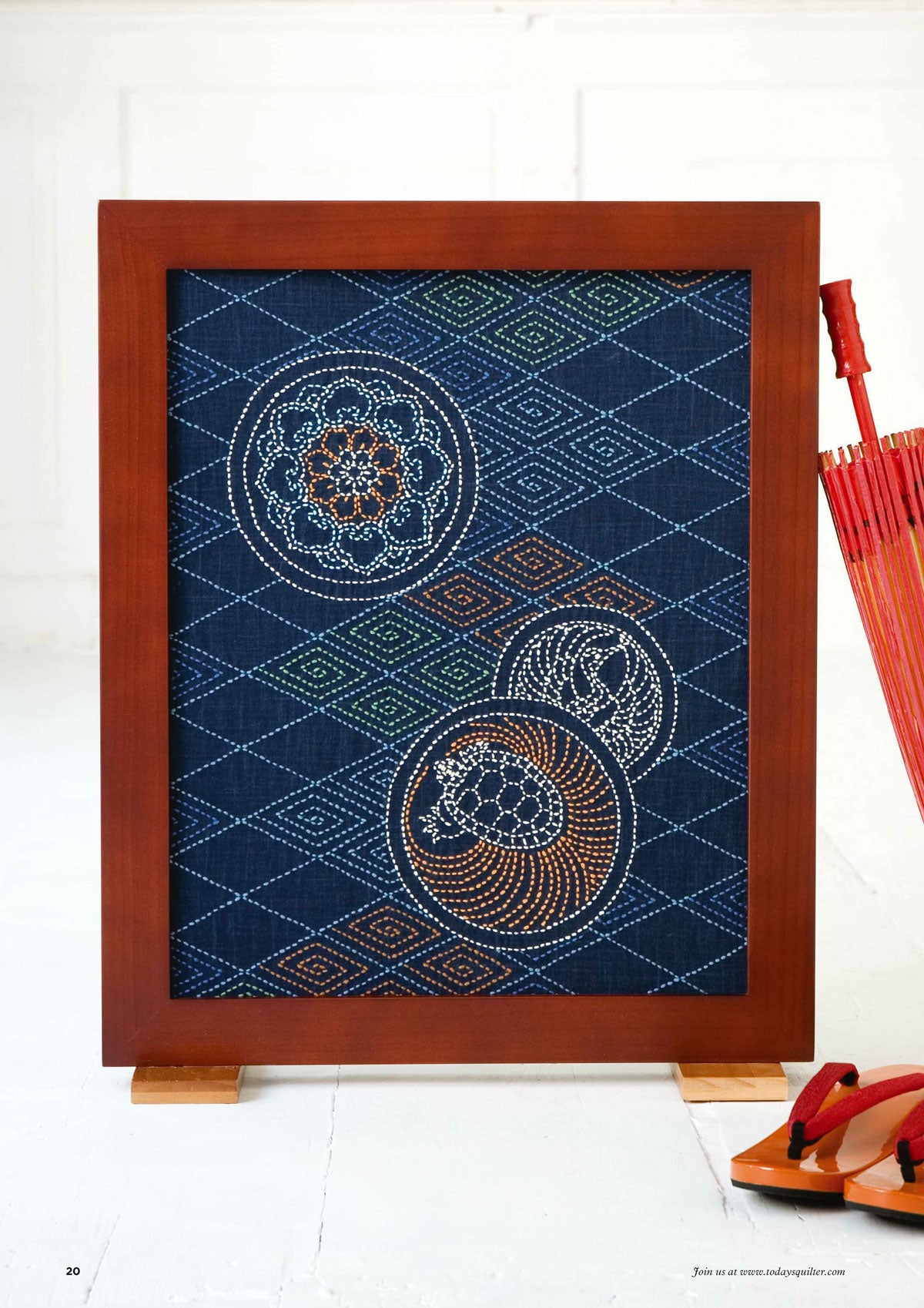 Framed textile art with geometric patterns and circular motifs in white on a dark blue background. A red umbrella and traditional sandals are nearby.