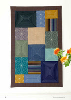 Patchwork quilt with geometric patterns in various colors, including blues, greens, and browns, hangs on a wall. Orange flowers in the foreground.