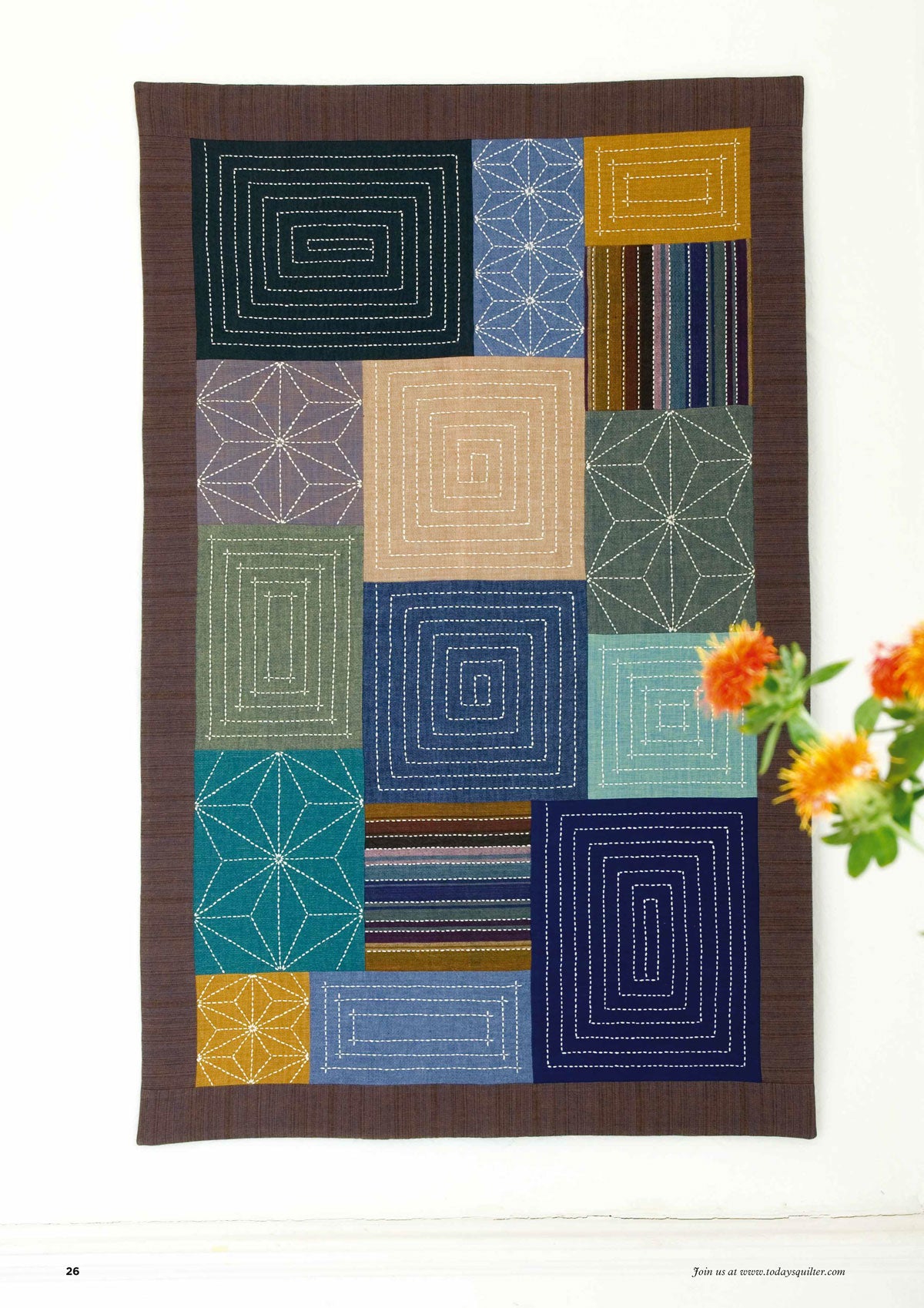Patchwork quilt with geometric patterns in various colors, including blues, greens, and browns, hangs on a wall. Orange flowers in the foreground.