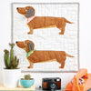 Quilt with dachshund design hanging on a wall with decor items below.