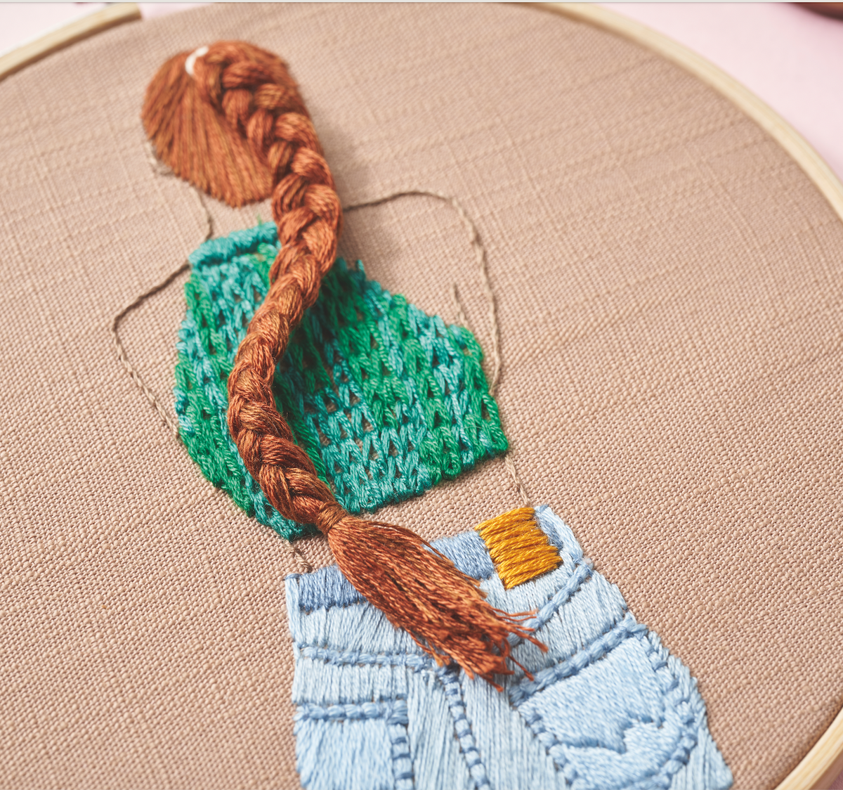 Embroidery hoop with a knitted green hat and braided brown hair on beige fabric.