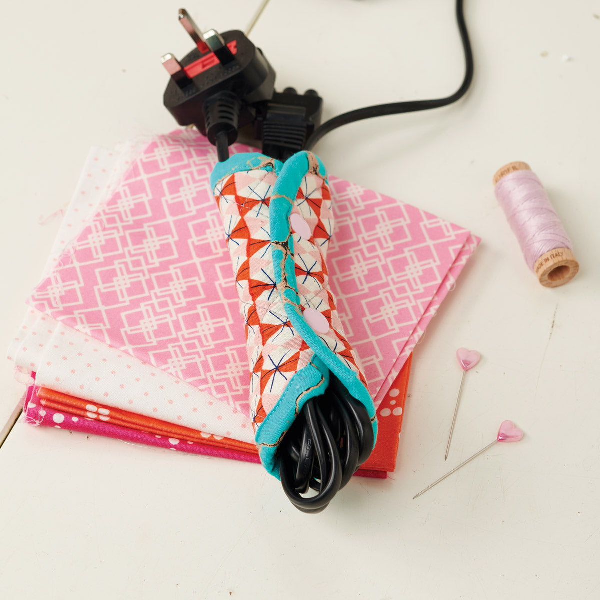 Fabric-wrapped electrical plug on a white surface with pink fabric and spools of thread.