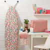 Ironing board with floral pattern next to a desk with fabric and stationery items against a white brick wall.