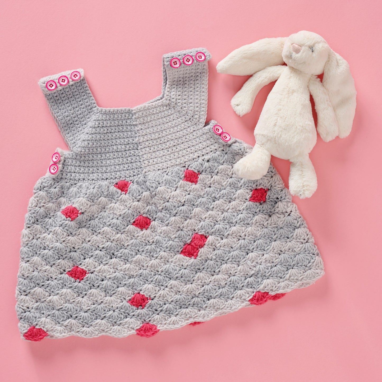 Crocheted baby dress with red flowers and a white plush bunny on a pink background