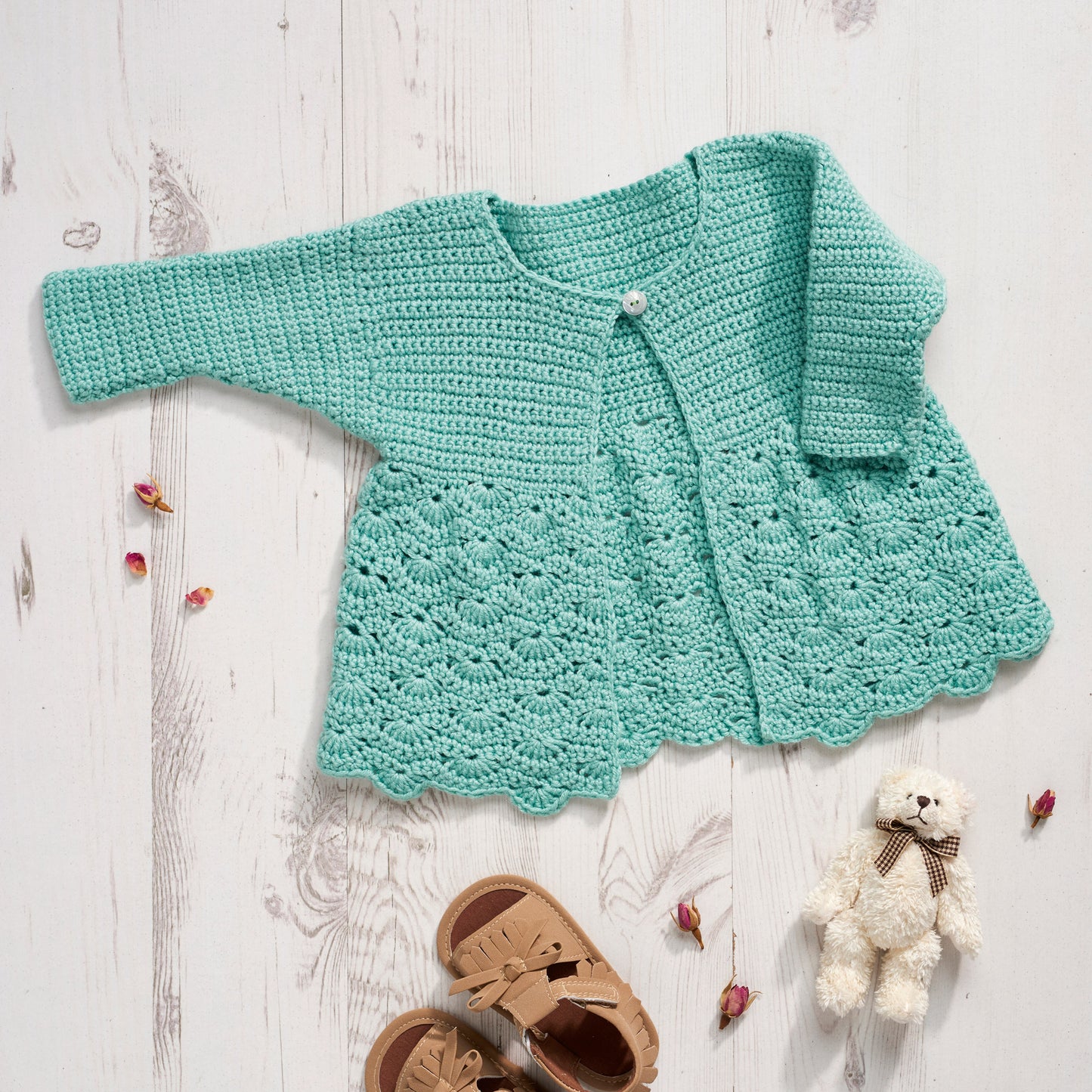 Crocheted turquoise baby cardigan on a light wooden surface with a teddy bear and sandals.