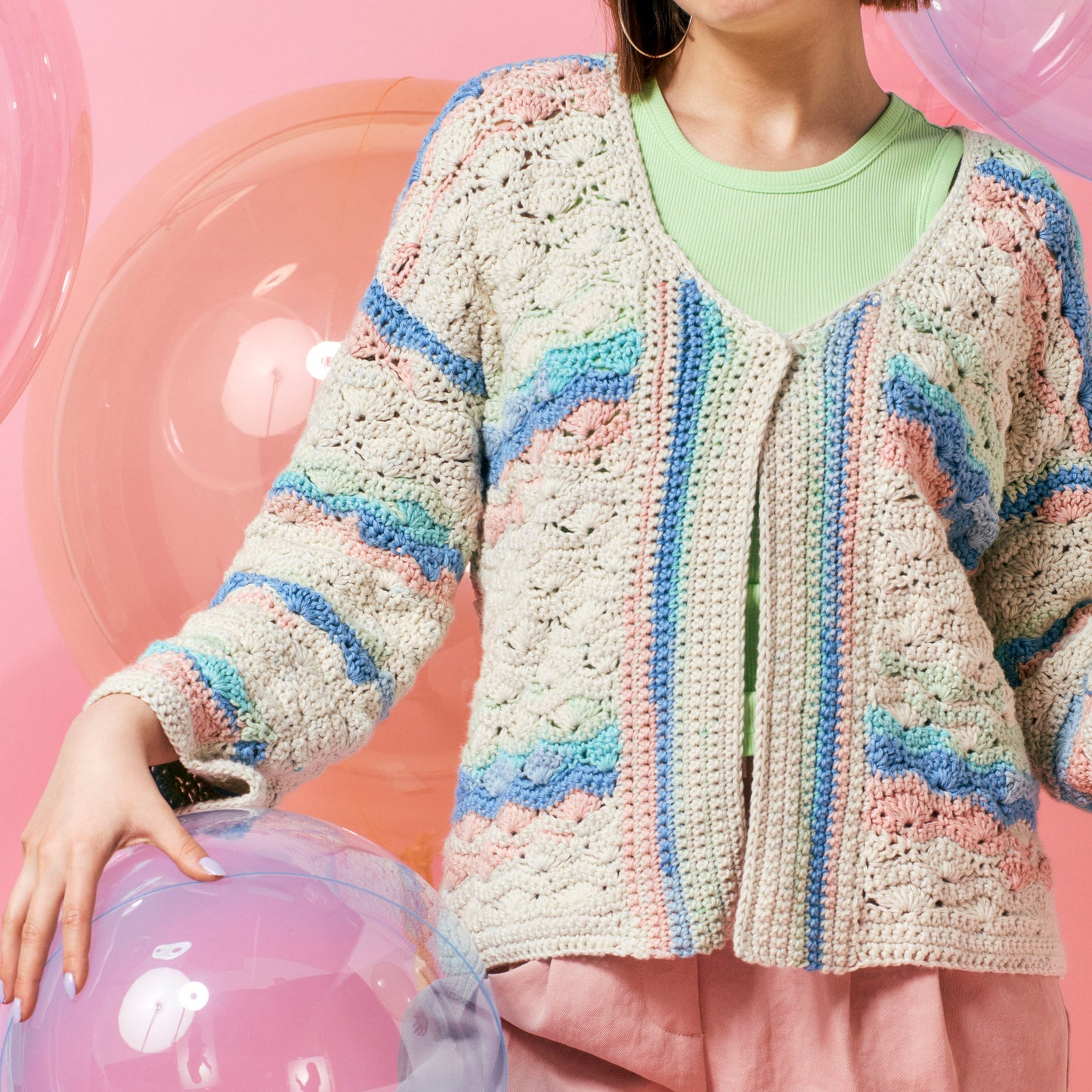A person wearing a pastel-colored crochet cardigan over a green shirt stands against a pink background. They are near large, iridescent bubbles, creating a playful, whimsical atmosphere.