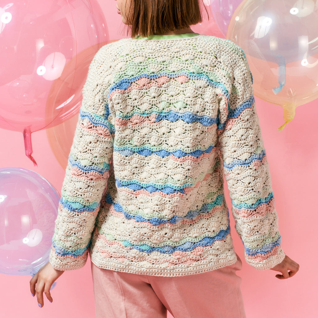 A person in a colorful crocheted sweater with pastel stripes stands against a pink background, holding translucent balloons, conveying a playful tone.