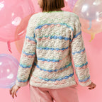A person in a colorful crocheted sweater with pastel stripes stands against a pink background, holding translucent balloons, conveying a playful tone.