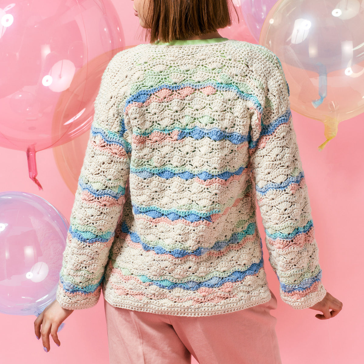 A person in a colorful crocheted sweater with pastel stripes stands against a pink background, holding translucent balloons, conveying a playful tone.
