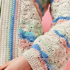 Close-up of a pastel crocheted sweater with intricate patterns in blue, pink, and green tones, set against a soft pink background. Cozy and textured.