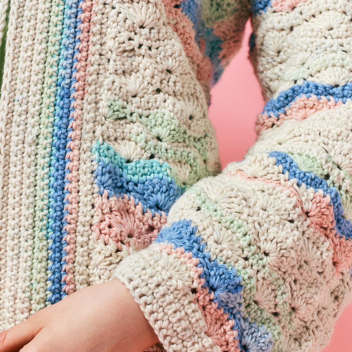 Close-up of a pastel crocheted sweater with intricate patterns in blue, pink, and green tones, set against a soft pink background. Cozy and textured.