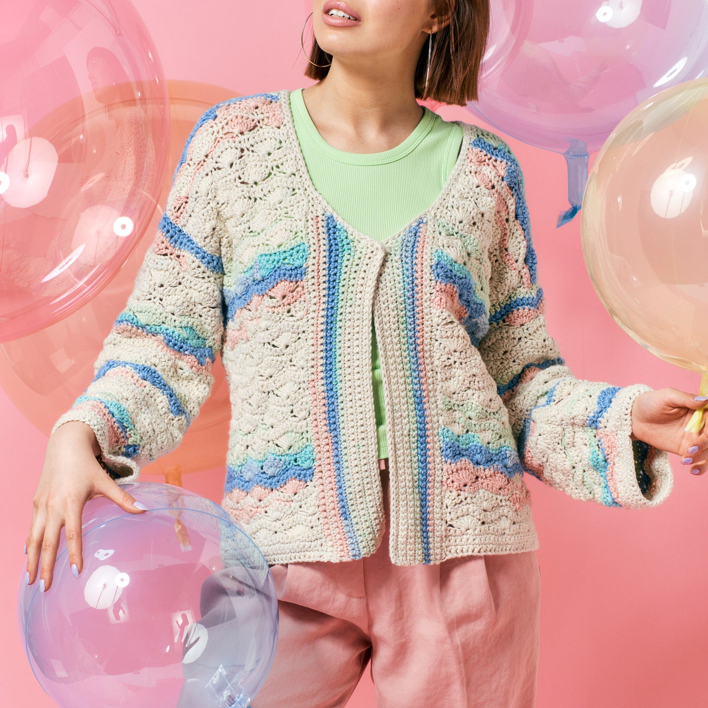 A person in pastel pink pants and a crochet cardigan with blue, pink, and green stripes. They're holding large transparent balloons against a pink backdrop.