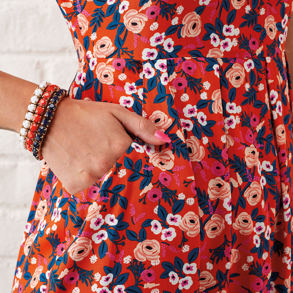 Close-up of a hand casually placed in the pocket of a vibrant, floral dress. The fabric is orange with pink and white flowers. The person wears multiple bracelets.