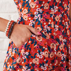 Close-up of a hand casually placed in the pocket of a vibrant, floral dress. The fabric is orange with pink and white flowers. The person wears multiple bracelets.