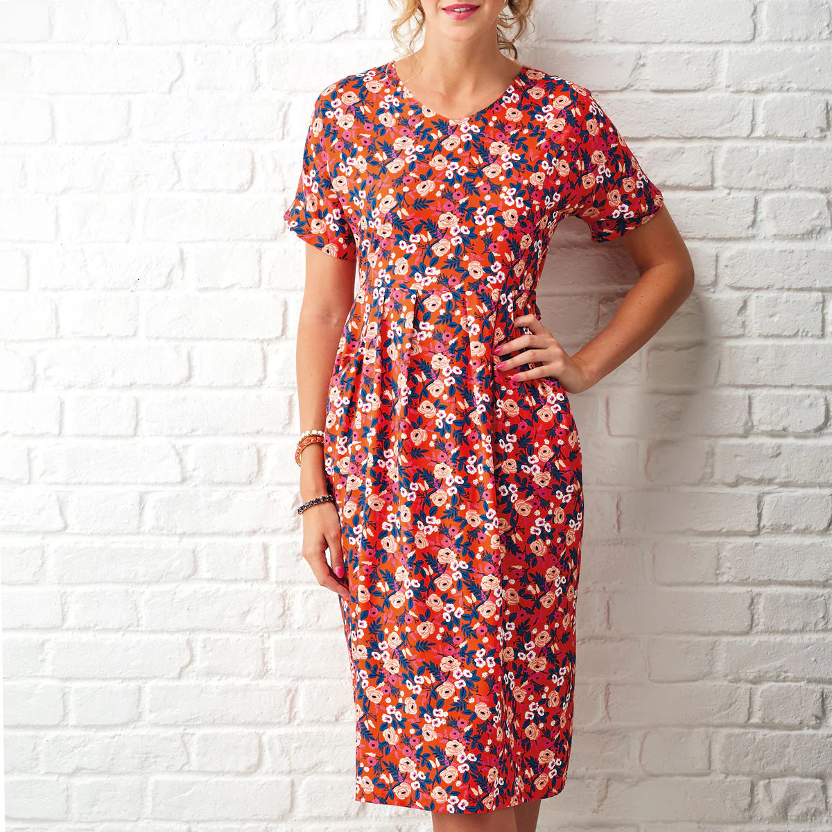 Woman wearing a colorful floral dress against a white brick wall.
